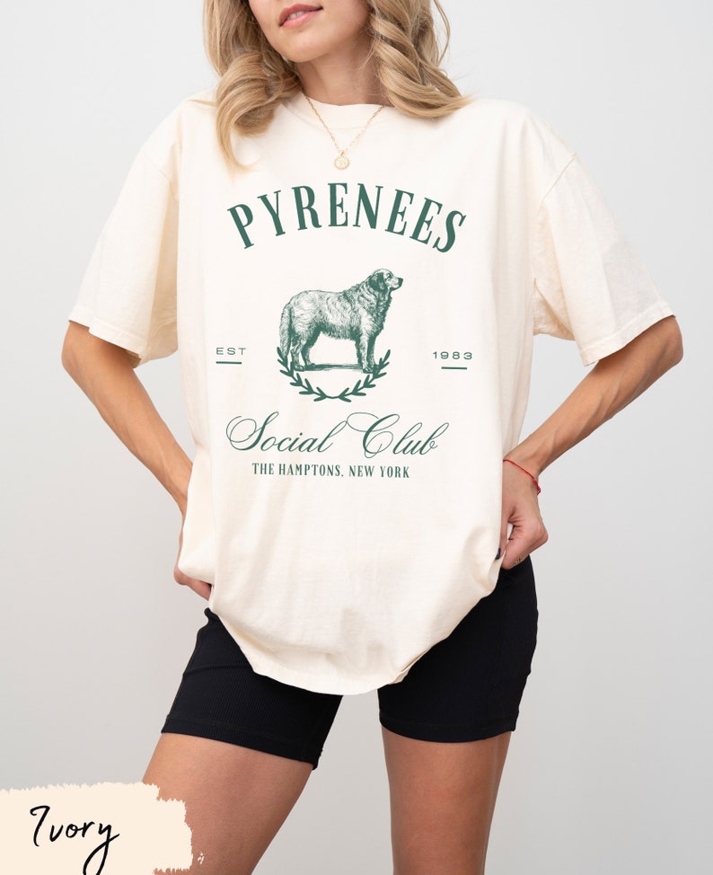May include: A cream-colored t-shirt with a green graphic of a dog and the text "Pyrenees Social Club The Hamptons, New York" printed on the front.