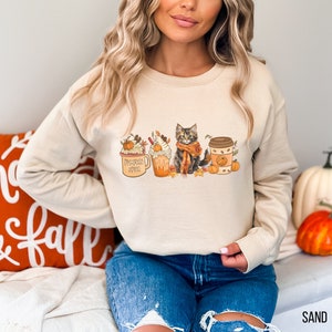 May include: A beige sweatshirt with a graphic of a cat, pumpkin spice latte, and a coffee cup. The graphic is in fall colors and features pumpkins and leaves. The text on the mug reads "Pumpkin Spice".