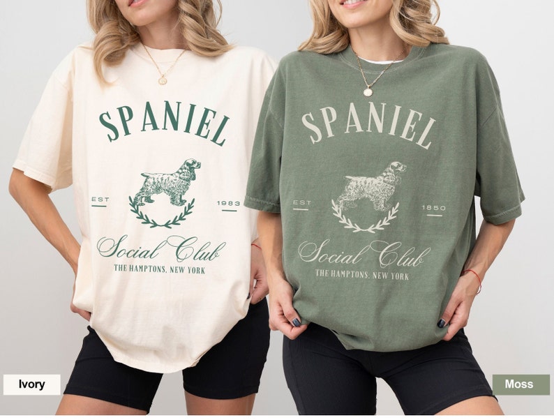 May include: Two women wearing oversized t-shirts with a vintage-style graphic of a spaniel dog and the text "SPANIEL Social Club THE HAMPTONS, NEW YORK". The t-shirt on the left is ivory and the t-shirt on the right is moss green.