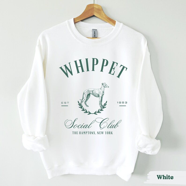 Puede incluir: Sudadera blanca con un dise&ntilde;o gr&aacute;fico verde que dice "Whippet Social Club" con un dibujo de un perro whippet y el texto "Est. 1983" y "The Hamptons, New York".