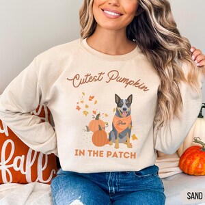 May include: Sand-colored sweatshirt with the text "Cutest Pumpkin IN THE PATCH". The design features a dog wearing an orange bandana, pumpkins, and autumn leaves. The word "Blue" is on the bandana.