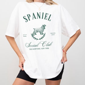 May include: White oversized t-shirt with a green graphic print of a spaniel dog, the text "SPANIEL", "EST 1983", "Social Club", and "THE HAMPTONS, NEW YORK".