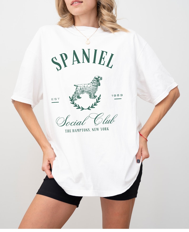May include: White oversized t-shirt with a green graphic print of a spaniel dog, the text "SPANIEL", "EST 1983", "Social Club", and "THE HAMPTONS, NEW YORK".