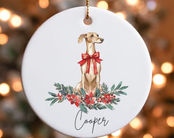 May include: A white ceramic Christmas ornament featuring a brown and white Italian Greyhound dog wearing a red bow. The dog is sitting in a wreath of green leaves and red flowers. The ornament is personalized with the name "Cooper".