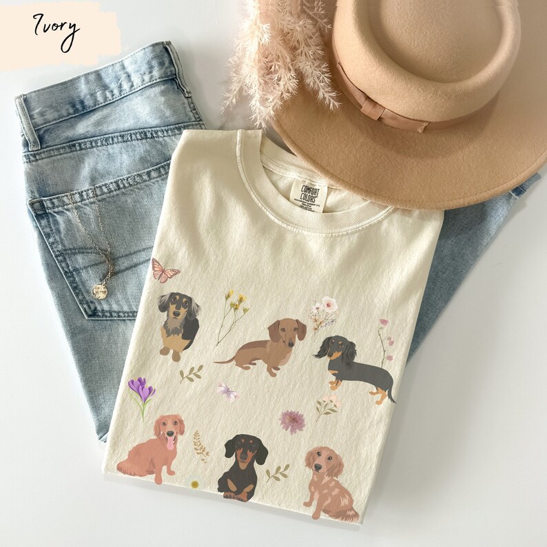 以下が含まれることがあります： アイボリーカラーのTシャツに、複数のダックスフントの犬と花のアクセントが描かれたグラフィックがプリントされています。Tシャツのタグには「COMFORT COLORS」の文字が見えます。