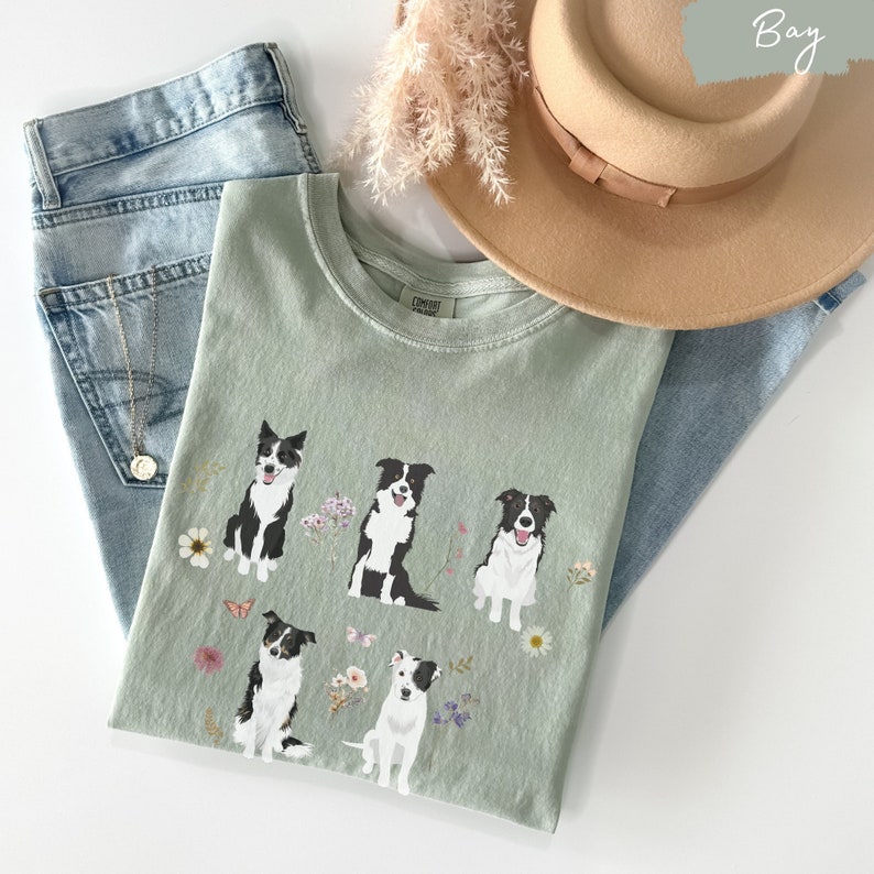 May include: A light green t-shirt with a graphic of four black and white border collies surrounded by flowers and butterflies. The shirt has a crew neck and short sleeves.