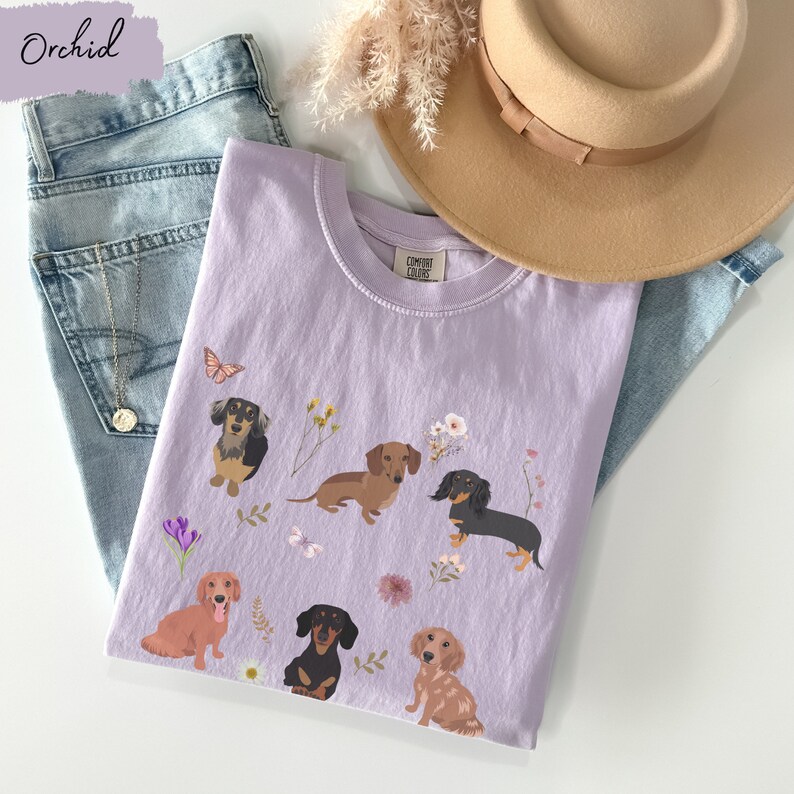 以下が含まれることがあります： ラベンダー色のTシャツに、ダックスフント5匹と花のアクセントが描かれたグラフィックがプリントされています。Tシャツには「COMFORT COLORS」というタグが付いています。