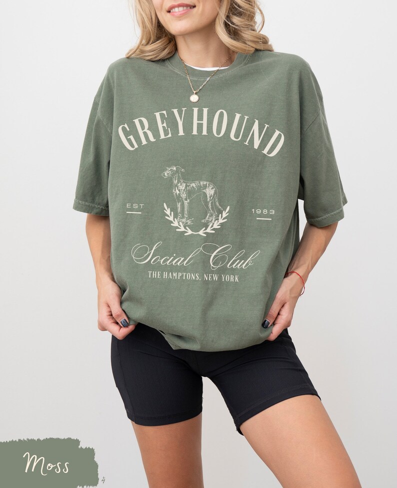 以下が含まれることがあります： モスグリーンのオーバーサイズTシャツに、ヴィンテージ風のグラフィックデザインが施されています。グラフィックには、グレイハウンドの犬、テキスト「Greyhound Social Club」、日付「EST 1983」が描かれています。