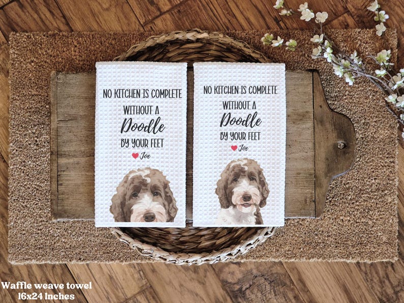 two white towels with a doodle of a dog's face and text printed on them, placed on a woven basket on a wooden surface.