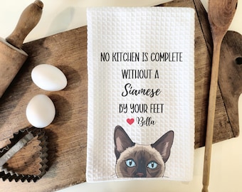 May include: White kitchen towel with the text "No kitchen is complete without a Siamese by your feet Bella" and a photo of a Siamese cat.