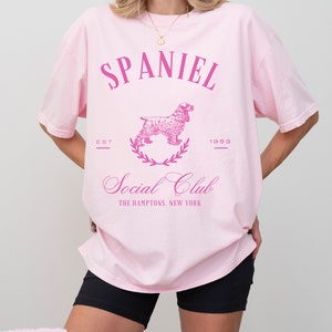 May include: Pink oversized t-shirt with a pink graphic of a spaniel dog, a wreath, and the text "SPANIEL EST 1983 Social Club THE HAMPTONS, NEW YORK"