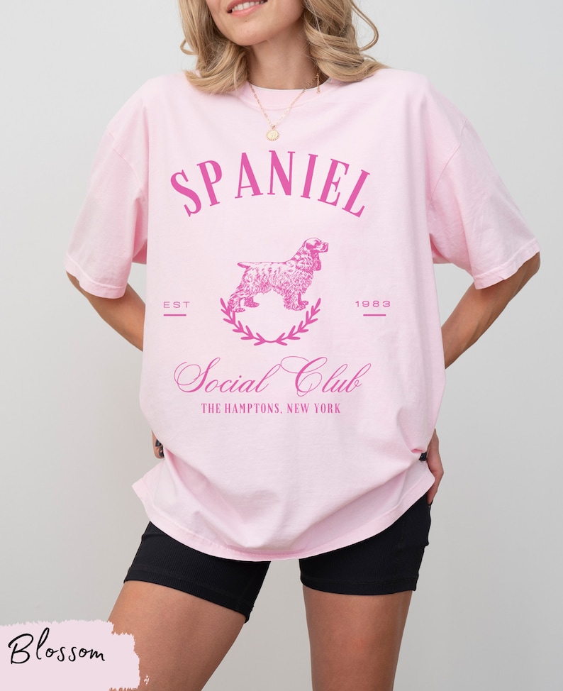 May include: Pink oversized t-shirt with a pink graphic of a spaniel dog, a wreath, and the text "SPANIEL EST 1983 Social Club THE HAMPTONS, NEW YORK"