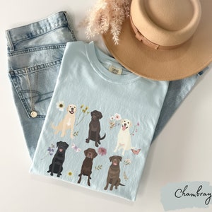 May include: A light blue t-shirt with a floral design and six Labrador Retriever dogs in different colors. The dogs are sitting and standing in a variety of poses. The shirt has the text "Comfort Colors" on the tag.