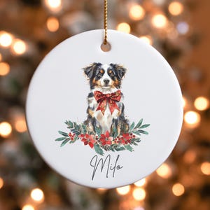 May include: A white ceramic Christmas ornament featuring a watercolor painting of a black, brown, and white dog wearing a red bow. The dog is surrounded by green leaves and red berries. The ornament is personalized with the name "Milo".