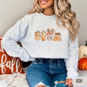 May include: A gray sweatshirt with a graphic design featuring a small dog wearing a scarf, a pumpkin spice latte, and a cup of coffee. The graphic is surrounded by fall-themed elements like leaves and pumpkins.