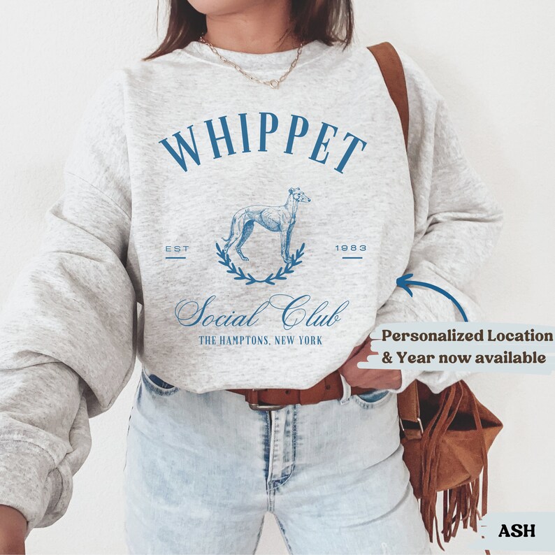 Puede incluir: Una sudadera gris con un dise&ntilde;o gr&aacute;fico azul que presenta un perro Whippet, el texto "Whippet Social Club" y los a&ntilde;os "EST 1983". El texto "The Hamptons, New York" tambi&eacute;n est&aacute; incluido en el dise&ntilde;o. La sudadera est&aacute; siendo usada por una persona. El texto "Ubicaci&oacute;n personalizada y a&ntilde;o ahora disponible" est&aacute; escrito en un cuadro blanco con un borde azul.