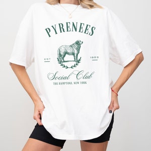 May include: White oversized t-shirt with a green graphic print of a dog, a wreath, and the text "Pyrenees Est 1983 Social Club The Hamptons, New York"