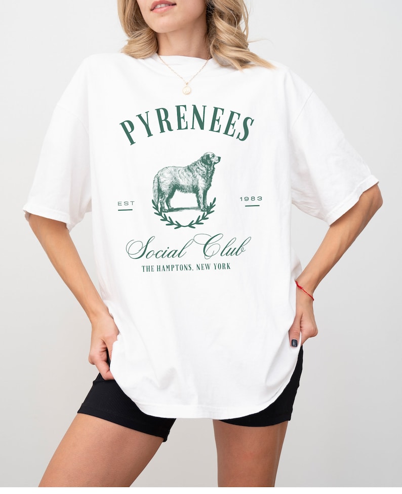 May include: White oversized t-shirt with a green graphic print of a dog, a wreath, and the text "Pyrenees Est 1983 Social Club The Hamptons, New York"