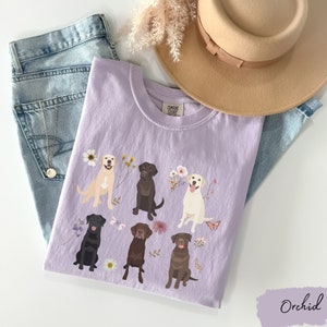 May include: A lavender t-shirt with a floral design featuring six Labrador Retrievers in different colors. The shirt has the text "COMFORT COLORS" on the tag.