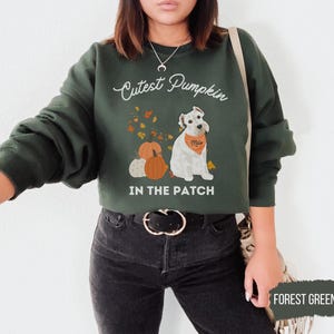 May include: Forest green sweatshirt with the text "Cutest Pumpkin In The Patch" and a graphic of a white dog wearing a bandana, with pumpkins and fall leaves. The sweatshirt is a crew neck style.