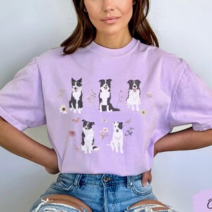 May include: A lavender t-shirt with a graphic of five black and white border collies sitting in a field of flowers. The shirt has a crew neck and short sleeves.