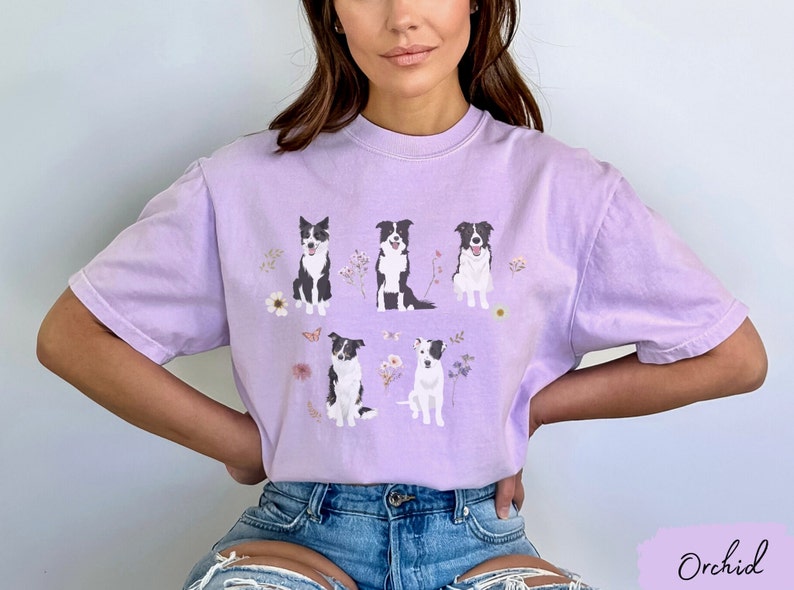 May include: A lavender t-shirt with a graphic of five black and white border collies sitting in a field of flowers. The shirt has a crew neck and short sleeves.