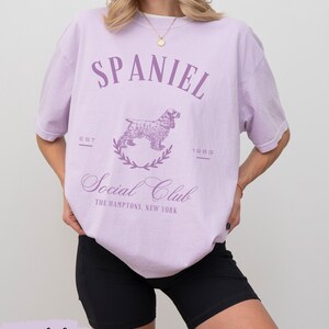 May include: A lavender t-shirt with a graphic of a spaniel dog and the text "Spaniel Social Club The Hamptons, New York" in purple. The text "Est 1983" is also printed on the shirt.