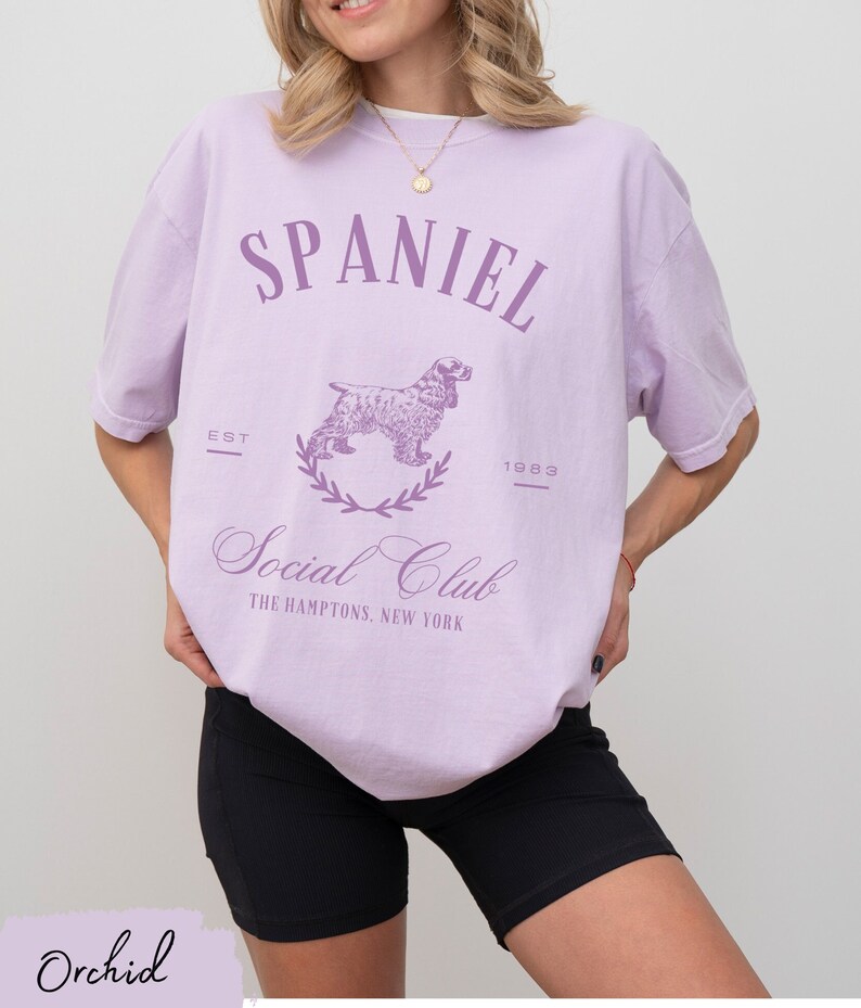 May include: A lavender t-shirt with a graphic of a spaniel dog and the text "Spaniel Social Club The Hamptons, New York" in purple. The text "Est 1983" is also printed on the shirt.
