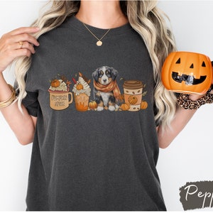 May include: A dark gray t-shirt with a graphic of a Bernese Mountain Dog puppy wearing a scarf, surrounded by fall-themed coffee cups and pumpkins. The text on one of the cups reads "Pumpkin Spice".