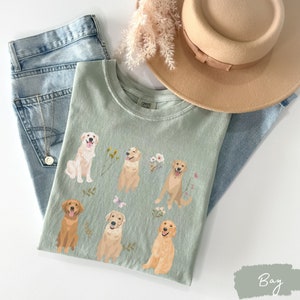 May include: A light green t-shirt with a graphic print of six golden retriever dogs with flowers and butterflies. The shirt is laid out on a pair of blue jeans and a straw hat.