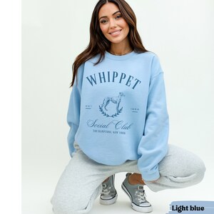 Puede incluir: Sudadera azul claro con un gr&aacute;fico de un perro whippet y el texto "Whippet Social Club The Hamptons, New York"