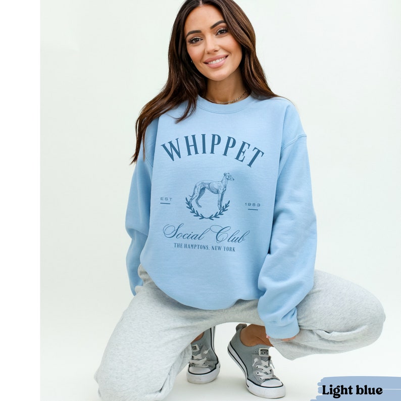 Puede incluir: Sudadera azul claro con un gr&aacute;fico de un perro whippet y el texto "Whippet Social Club The Hamptons, New York"