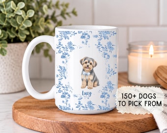 Puede incluir: Una taza de cerámica blanca con un patrón floral azul y blanco y una ilustración central de un perro pequeño con una pajarita azul claro. El nombre Lucy está impreso debajo del perro. Una etiqueta dice 150+ DOGS TO PICK FROM.