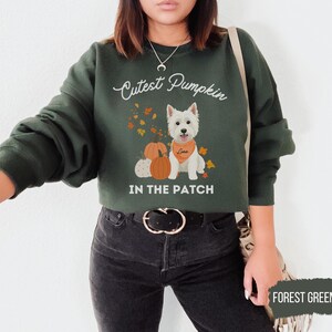 May include: Forest green sweatshirt with the text "Cutest Pumpkin In The Patch". The design features a white dog wearing an orange bandana, pumpkins, and autumn leaves. The sweatshirt is a crew neck style.