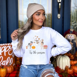 Puede incluir: Sudadera blanca con un gr&aacute;fico de tres perros, una calabaza y hojas de oto&ntilde;o. El texto "Tis the season" est&aacute; impreso debajo del gr&aacute;fico.