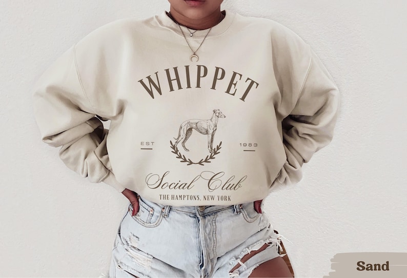 Puede incluir: Una sudadera beige con el texto "Whippet Social Club" y un gr&aacute;fico de un perro whippet. El texto "Est. 1983" est&aacute; debajo del gr&aacute;fico. La sudadera la lleva puesta una persona.