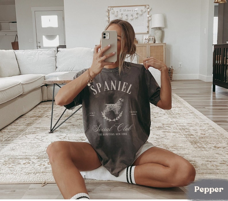 May include: A woman wearing a gray t-shirt with a graphic of a dog and the text "SPANIEL Social Club THE HAMPTONS, NEW YORK" printed on it. The shirt is a relaxed fit and has a distressed look. The woman is sitting on a rug with her legs crossed.