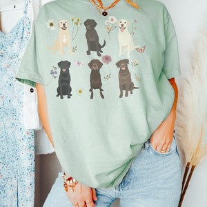 May include: A light green t-shirt with a graphic of six Labrador Retrievers in different colors, surrounded by flowers and butterflies.