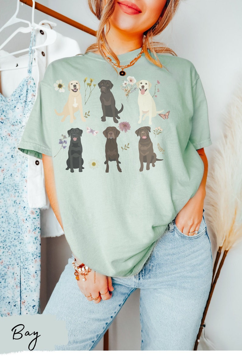 May include: A light green t-shirt with a graphic of six Labrador Retrievers in different colors, surrounded by flowers and butterflies.