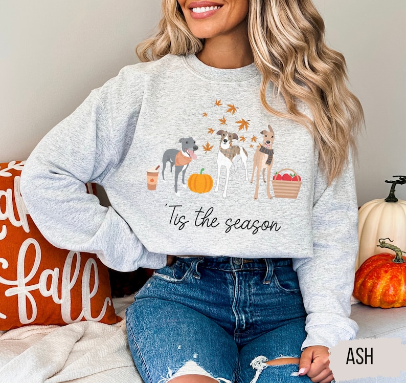 Puede incluir: Una sudadera gris con un gr&aacute;fico de tres perros, una calabaza y hojas de oto&ntilde;o. El texto "'Tis the season" est&aacute; impreso debajo del gr&aacute;fico.