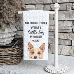 A white towel with a dog image and text hangs on a white towel rack next to a wicker basket and a brick wall.