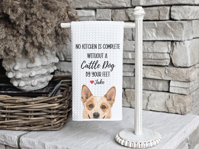 A white towel with a dog image and text hangs on a white towel rack next to a wicker basket and a brick wall.