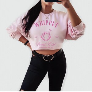 Puede incluir: Sudadera rosa recortada con el texto "Whippet Social Club" y un gr&aacute;fico de un perro whippet en una corona de laurel. El texto "Est. 1989" est&aacute; debajo del gr&aacute;fico. La sudadera la lleva puesta una persona con pantalones negros y un cintur&oacute;n dorado.