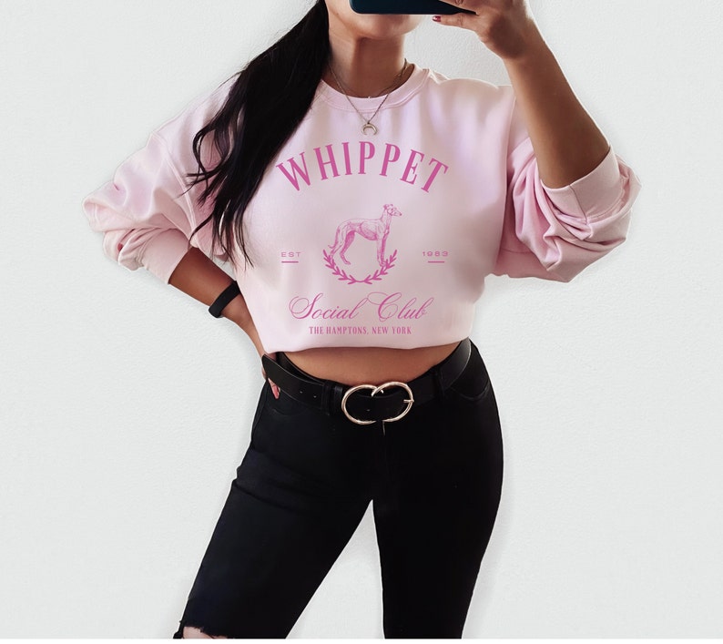Puede incluir: Sudadera rosa recortada con el texto "Whippet Social Club" y un gr&aacute;fico de un perro whippet en una corona de laurel. El texto "Est. 1989" est&aacute; debajo del gr&aacute;fico. La sudadera la lleva puesta una persona con pantalones negros y un cintur&oacute;n dorado.