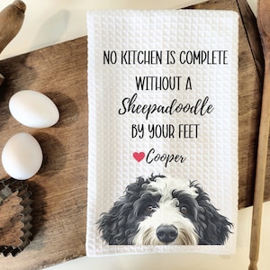 May include: White kitchen towel with a black and white Sheepadoodle dog peeking out from the bottom. The towel has the text "No kitchen is complete without a Sheepadoodle by your feet" and a red heart with the name "Cooper" underneath.