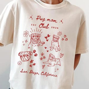 May include: Cream-colored t-shirt with a red graphic featuring pug illustrations, flowers, hearts, and stars. Text includes "Pug Mom Club EST 2026" and "San Diego, California".