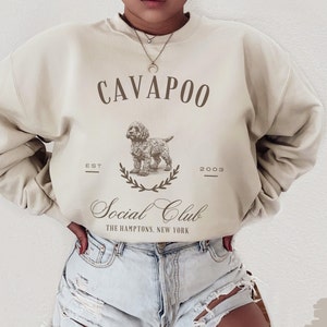 May include: A beige sweatshirt with the text "CAVAPOO Social Club THE HAMPTONS, NEW YORK" and a drawing of a dog. The text "EST 2003" is also on the sweatshirt.