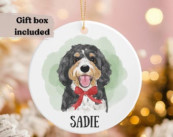 May include: A white ceramic ornament featuring a watercolor illustration of a dog with black, brown, and white fur, a red bow tie, and the name "Sadie" below. The background has a green watercolor wash and a gold string for hanging.