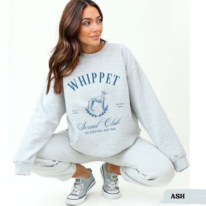 Puede incluir: Una sudadera gris con un estampado gr&aacute;fico azul de un perro whippet y el texto "Whippet Social Club The Hamptons, New York"  EST 1983.