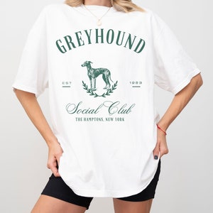 以下が含まれることがあります： グレーのグレイハウンド犬、月桂樹の輪、そして「Greyhound Social Club The Hamptons, New York」というテキストと「Est 1983」というテキストが書かれたグリーンのグラフィックプリントが施された、白のオーバーサイズTシャツです。